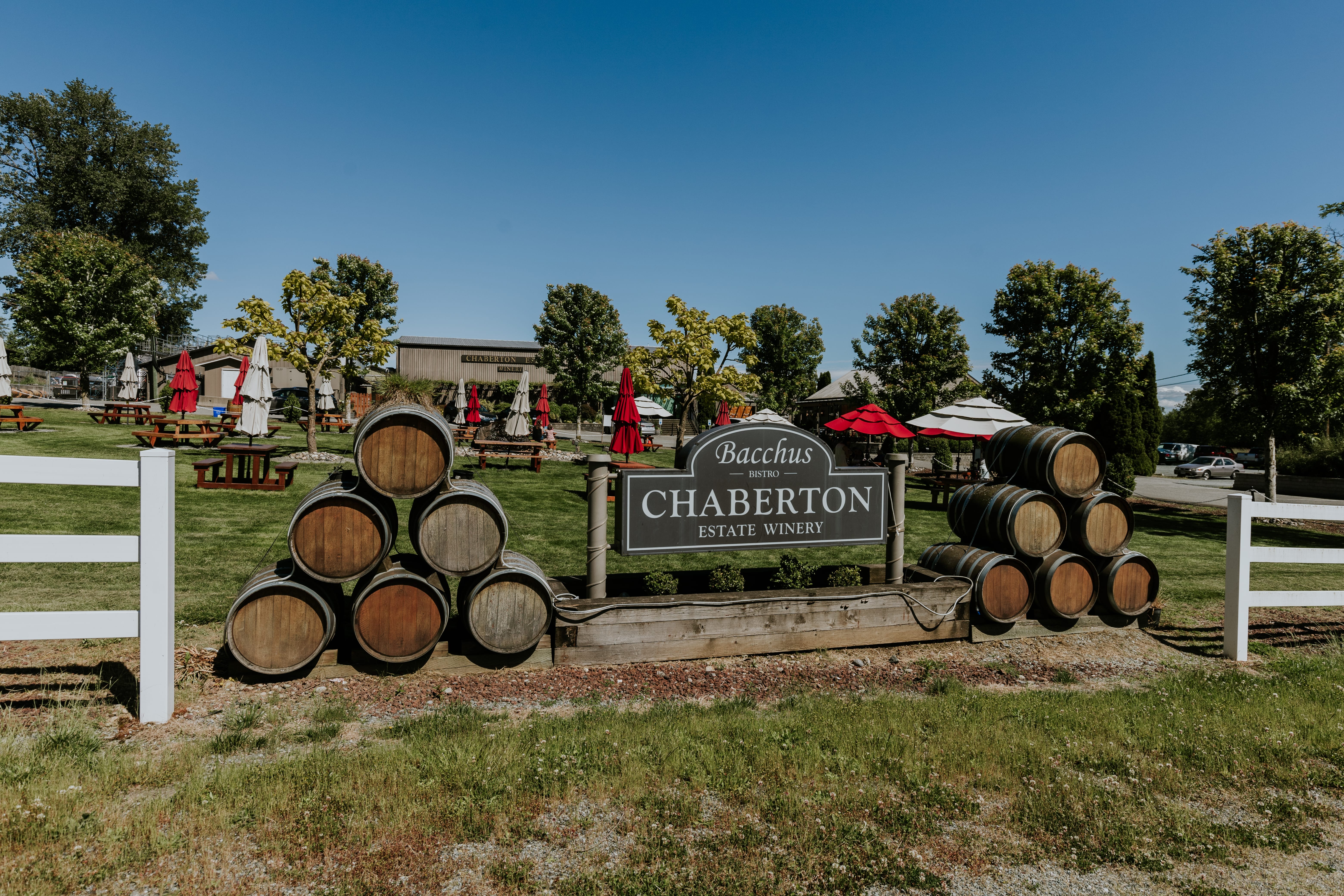 Entrance sign for Chaberton Estate Winery with stacked wine barrels on each side; outdoor seating with red umbrellas and trees in the background on a sunny day.