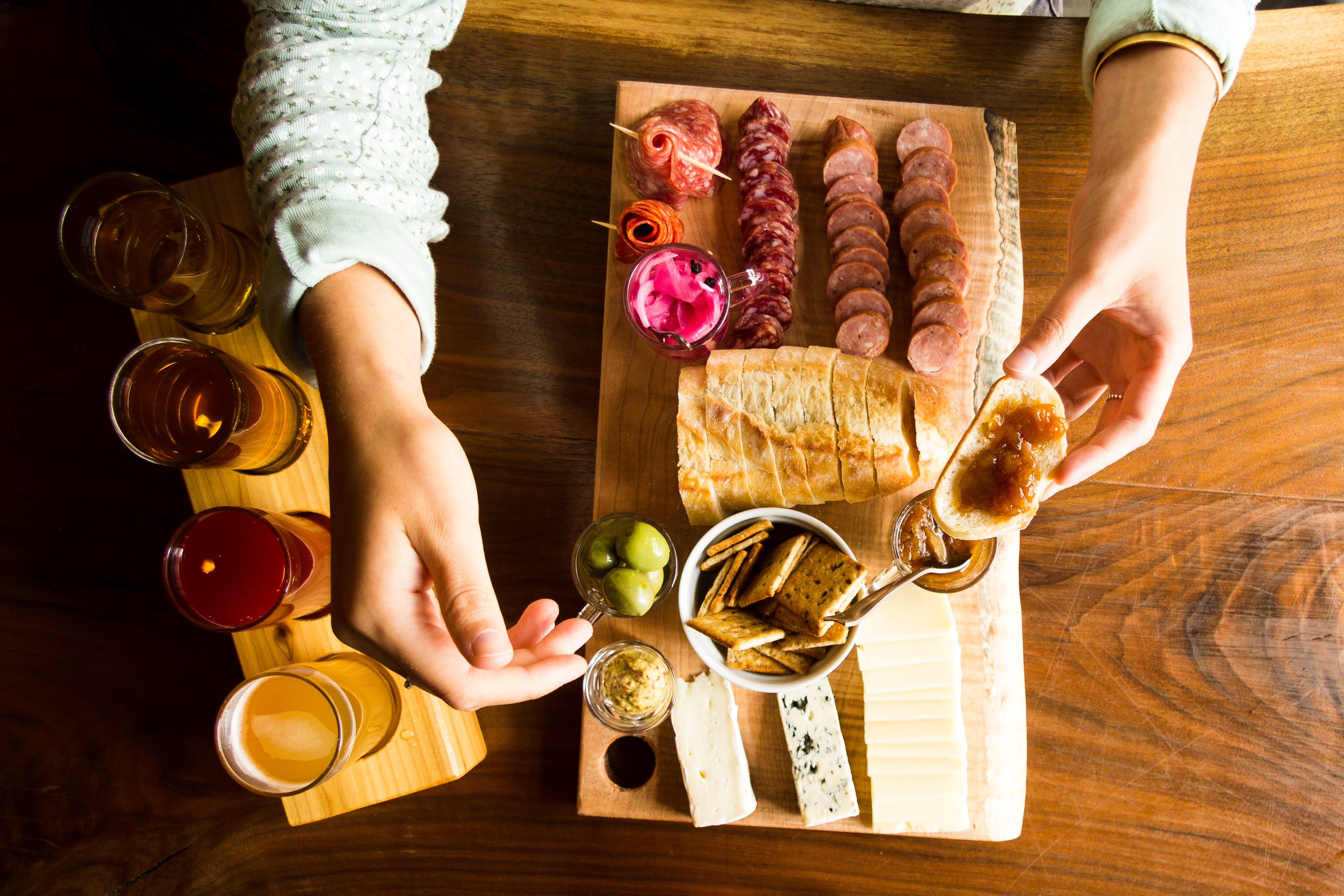A person sits at a table with a charcuterie board featuring assorted meats, cheeses, bread, crackers, pickles, and four glasses of beer, holding bread with a spread.