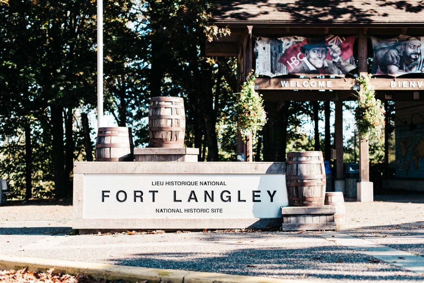 A sign reading "Fort Langley National Historic Site" is displayed in front of wooden barrels and a pavilion with welcome signs and murals.