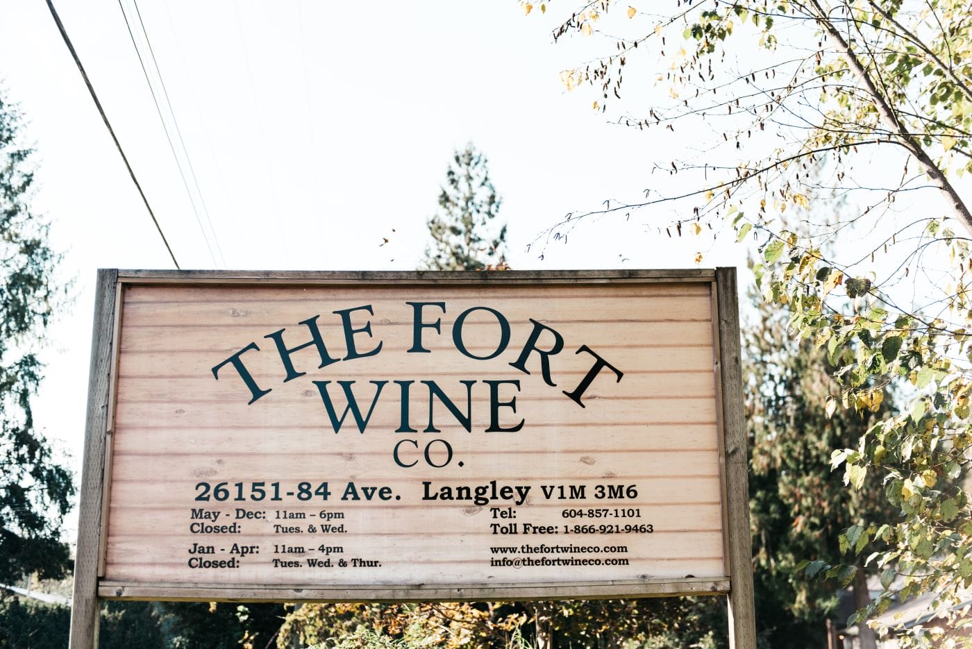 A wooden sign for The Fort Wine Co. shows address, hours, contact numbers, website, and email, surrounded by trees and power lines in the background.