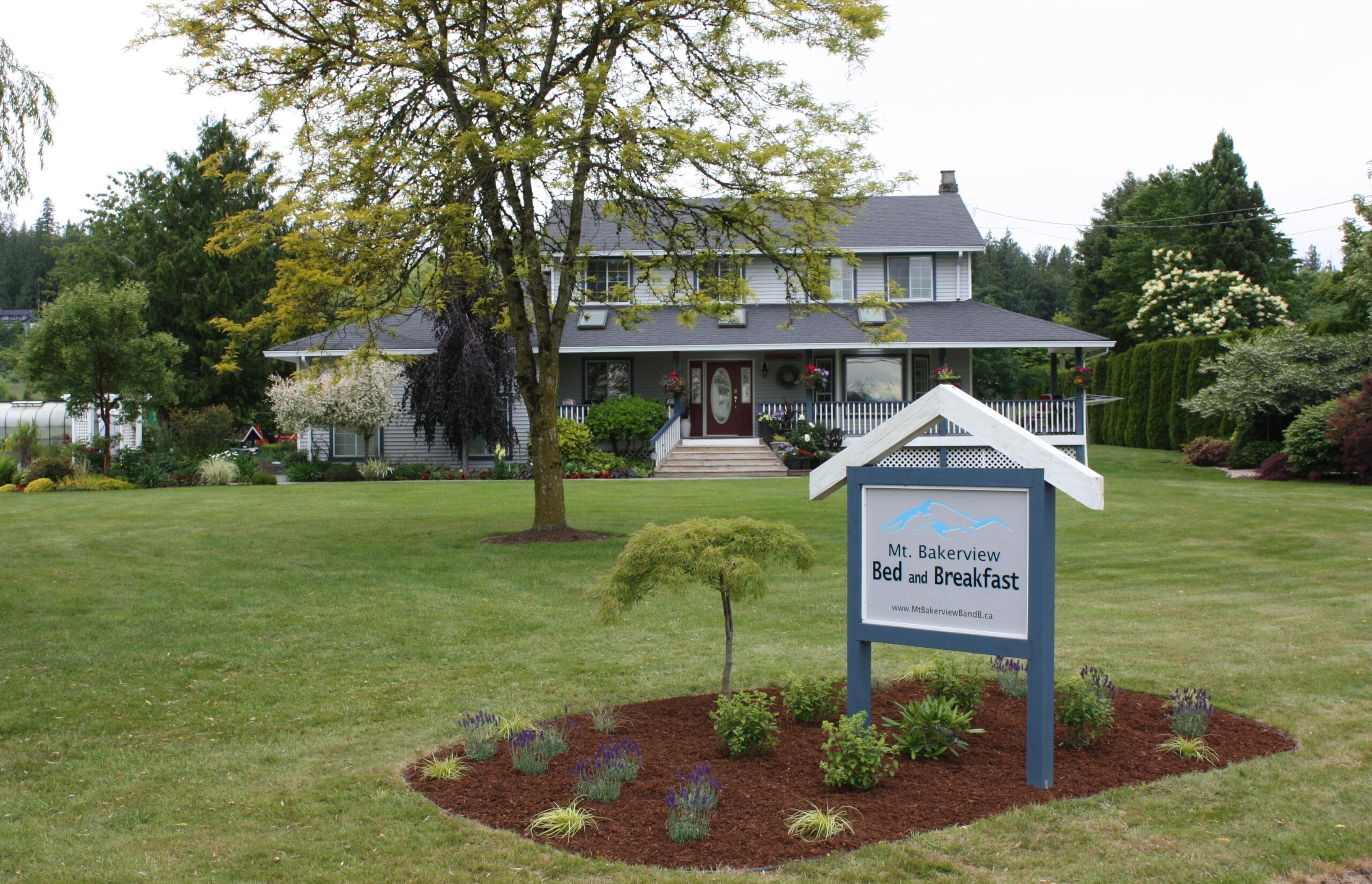A large house with a wraparound porch sits behind a landscaped yard and a sign reading "Mt. Bakerview Bed and Breakfast.