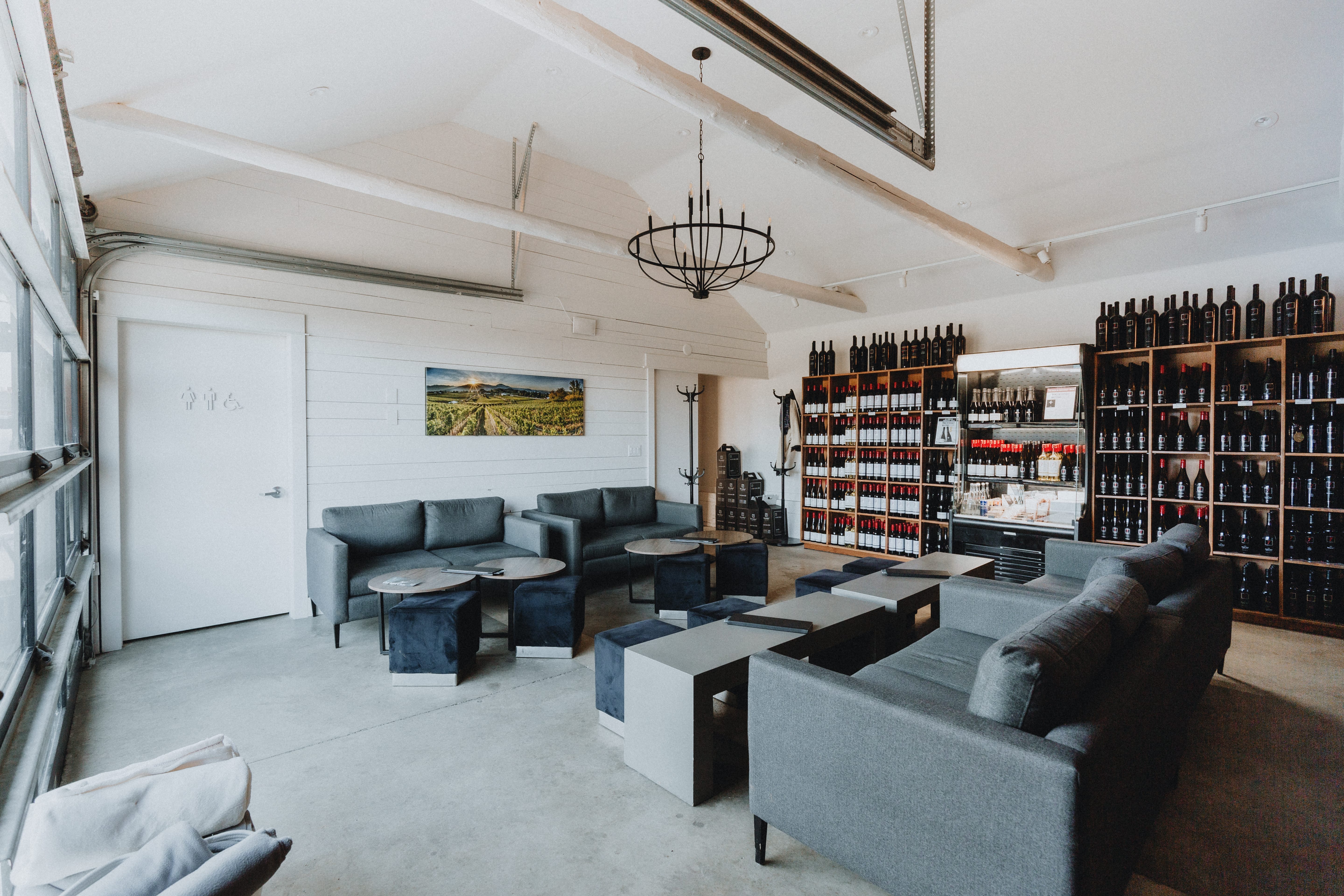 Modern tasting room with gray sofas, black cube tables, a wall of wine bottles, a chandelier, and a painted landscape on the wall. Natural light enters from large glass doors.