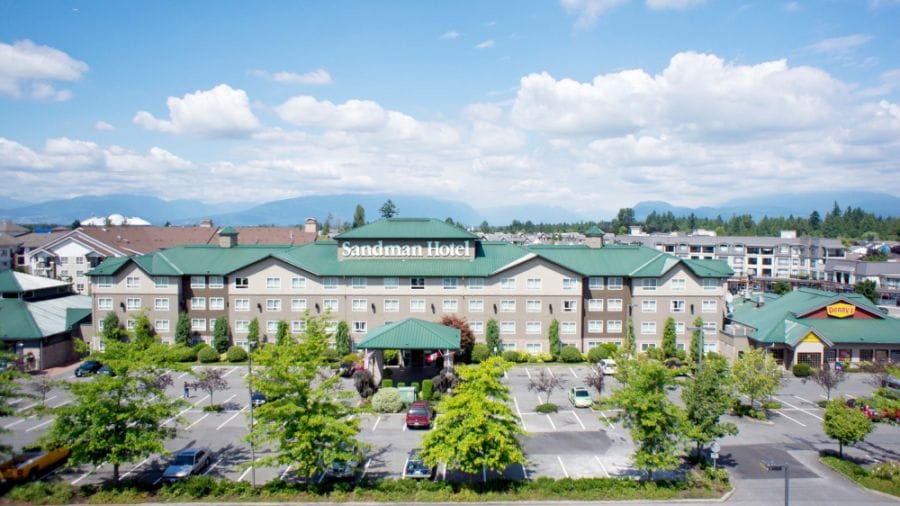A three-story Sandman Hotel with a green roof, surrounded by trees and a parking lot, on a sunny day with mountains visible in the background.