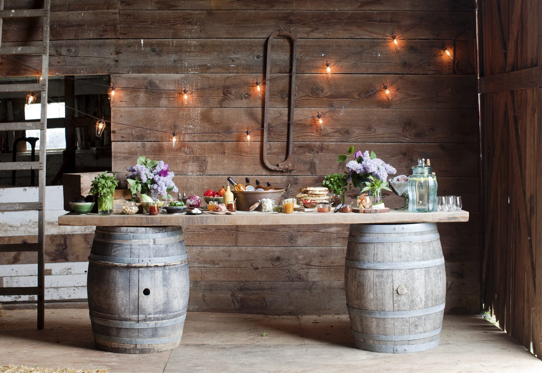 A rustic table made from two barrels and a wooden plank holds assorted food, drinks, flowers, and glassware in a barn with string lights on the wooden wall behind.