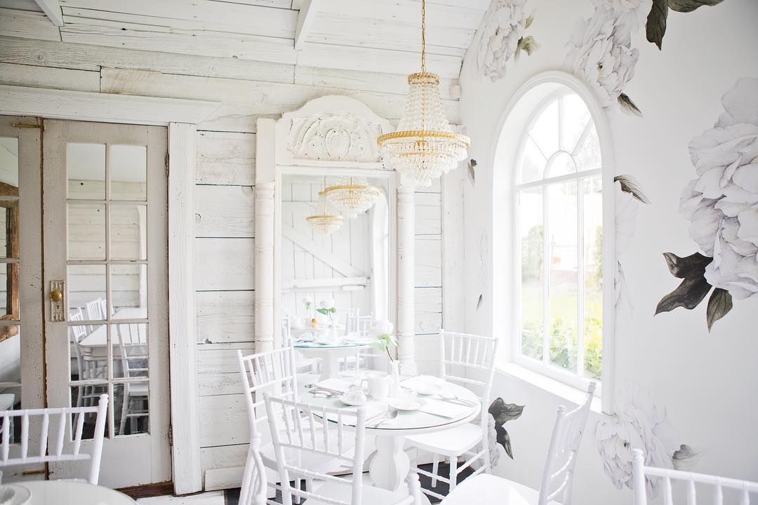 A bright, airy room with white wooden walls, round glass tables, white chairs, chandeliers, and large floral wallpaper near arched windows.