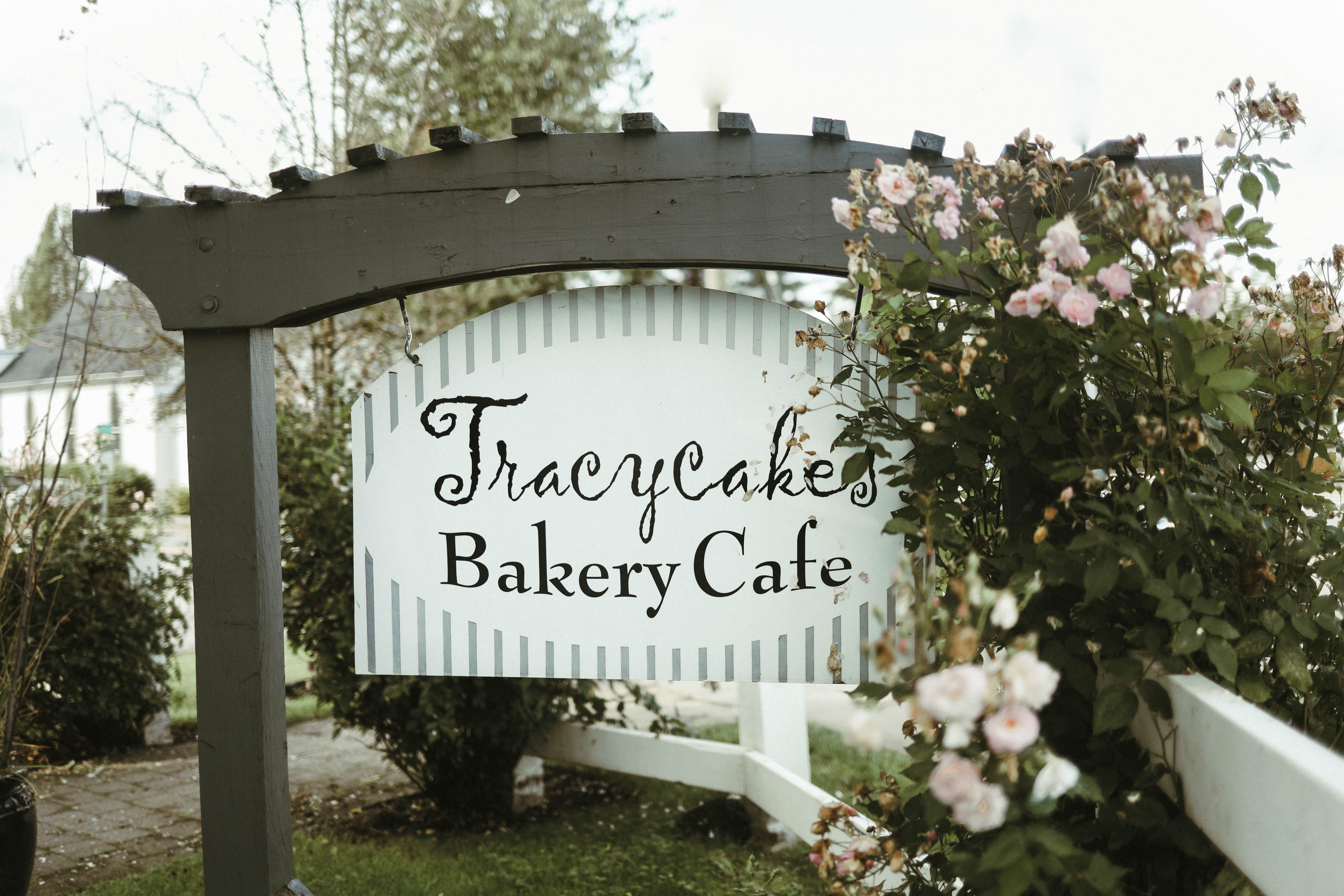 A wooden sign reading "Tracycakes Bakery Cafe" hangs from a dark pergola beside blooming pink flowers and green shrubbery.