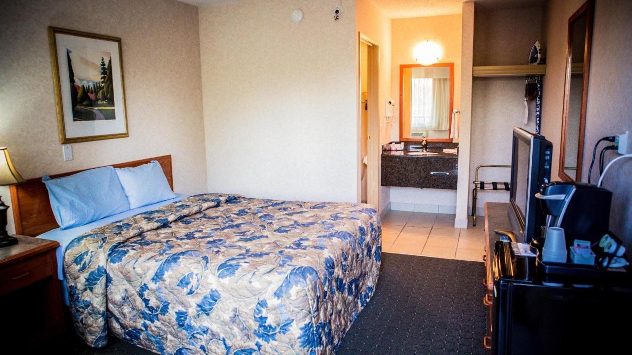 A hotel room with a bed, nightstand, wall art, TV, mini fridge, sink area, open closet, and a tiled area leading to a bathroom.