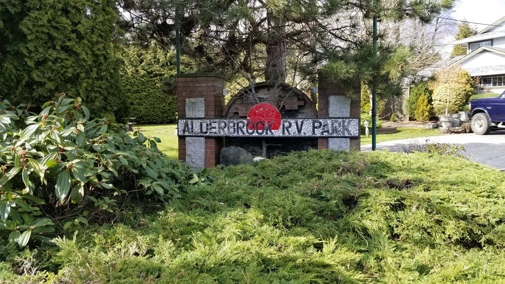 A weathered sign reading "Alderbrook RV Park" stands among greenery and shrubs, with a tree and a house visible in the background.