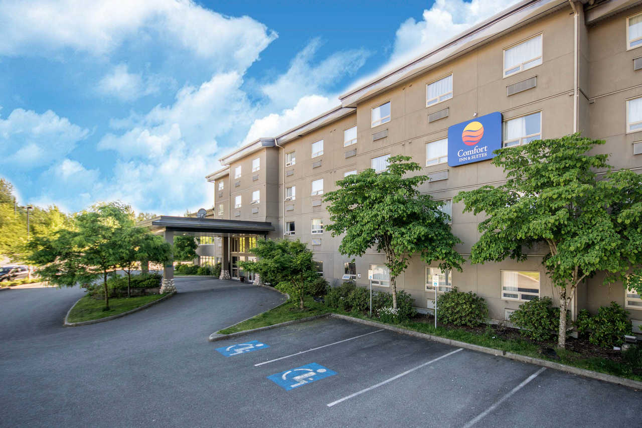 Four-story Comfort Inn & Suites hotel exterior with a driveway, trees, and two accessible parking spaces in front, under a partly cloudy sky.