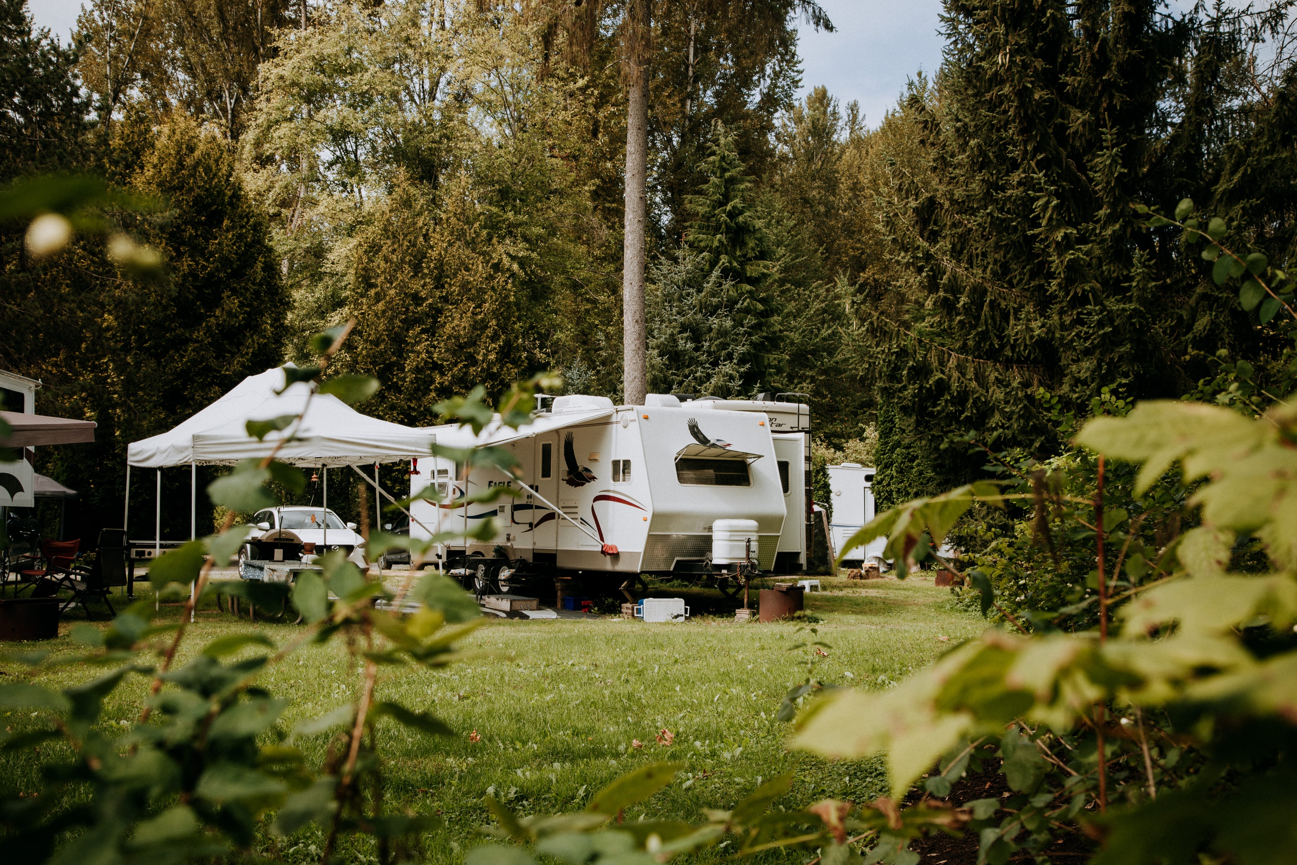 A white RV is parked on grass in a wooded campground, with a canopy, picnic table, and outdoor chairs nearby. Trees and greenery surround the area.