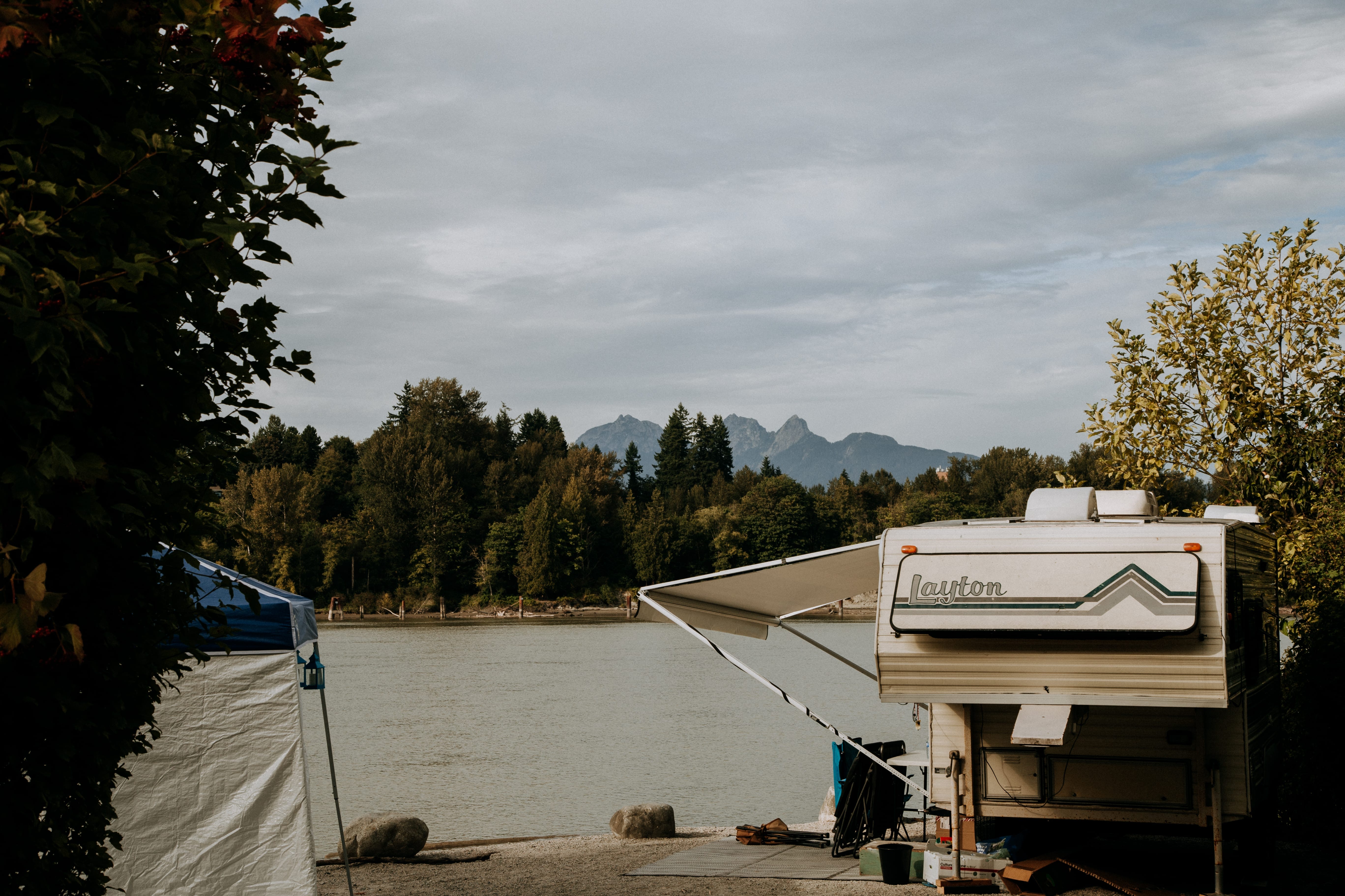 A camper with an extended awning is parked by a calm river, surrounded by trees, with mountains visible in the background under a cloudy sky.