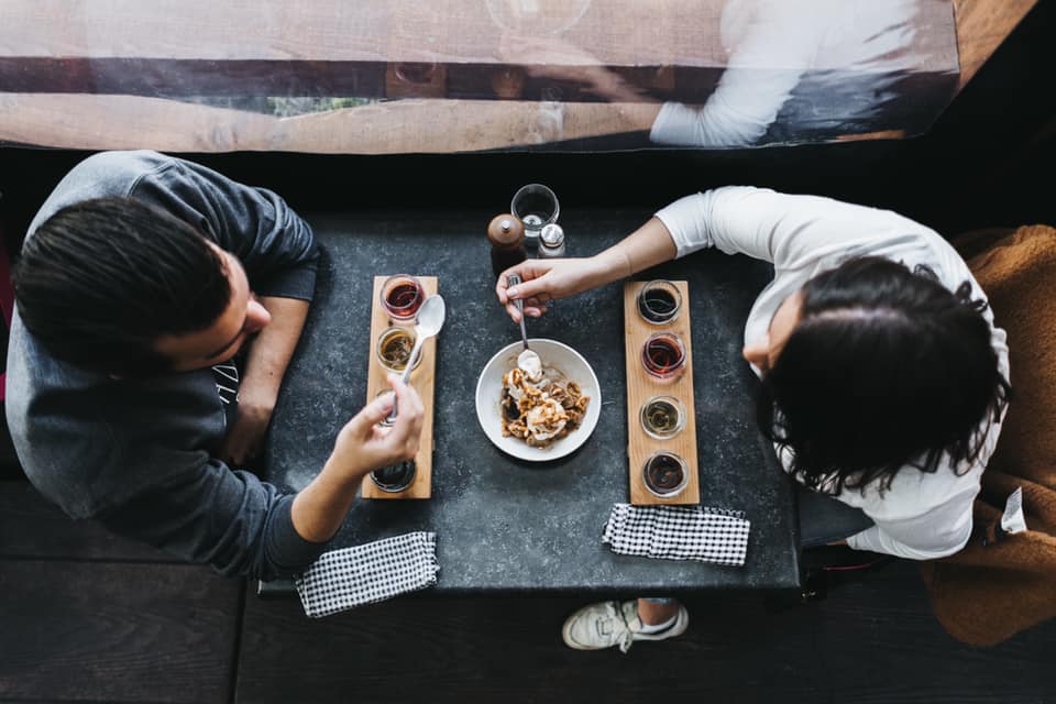 Two people sit across from each other at a table, sharing a dessert and sampling drinks served in small glasses on wooden trays.