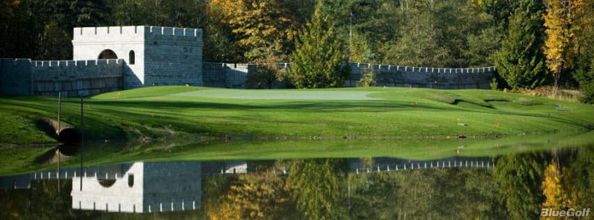 A golf course green with a stone wall and turret in the background, surrounded by trees and reflected in a pond in the foreground.