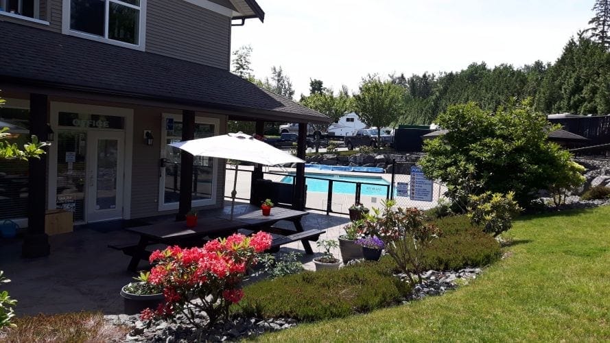 A picnic table with an umbrella sits on a patio next to a building, overlooking a landscaped garden and an outdoor swimming pool. Trees and RVs are visible in the background.