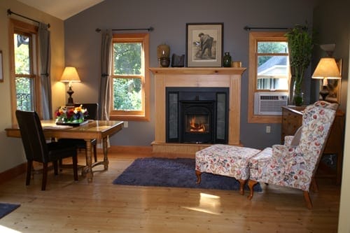 A cozy living room with a lit fireplace, floral armchair, small dining table, lamps, and windows letting in natural light.