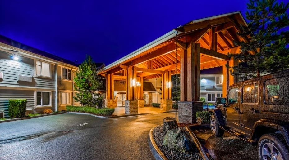 A well-lit hotel entrance with a large wooden canopy, surrounded by buildings and parked cars, at night.