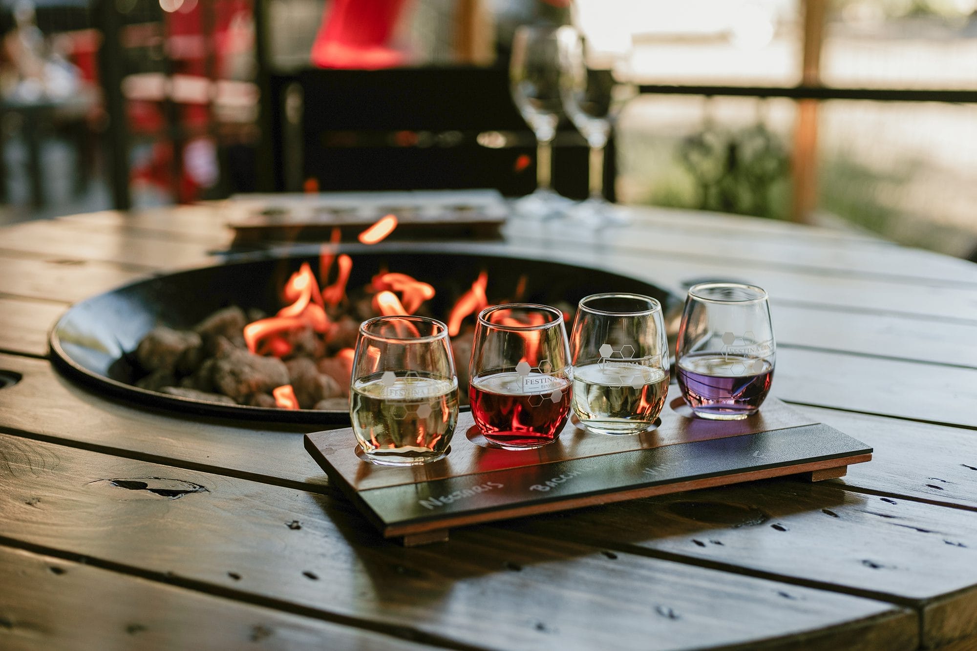 Four glasses of wine are arranged on a wooden tray atop a wooden table near a fire pit, with two empty wine glasses and chairs in the background.
