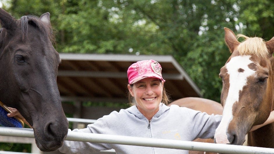 A woman in a pink cap and gray sweatshirt stands between two horses, smiling, with trees and a shelter in the background.