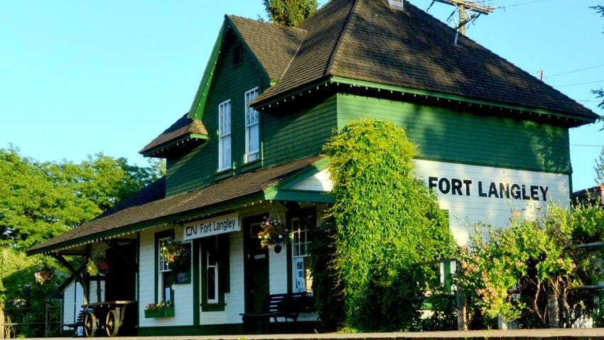 Green and white historic train station building labeled “Fort Langley,” with hanging flower baskets and dense greenery around the entrance.