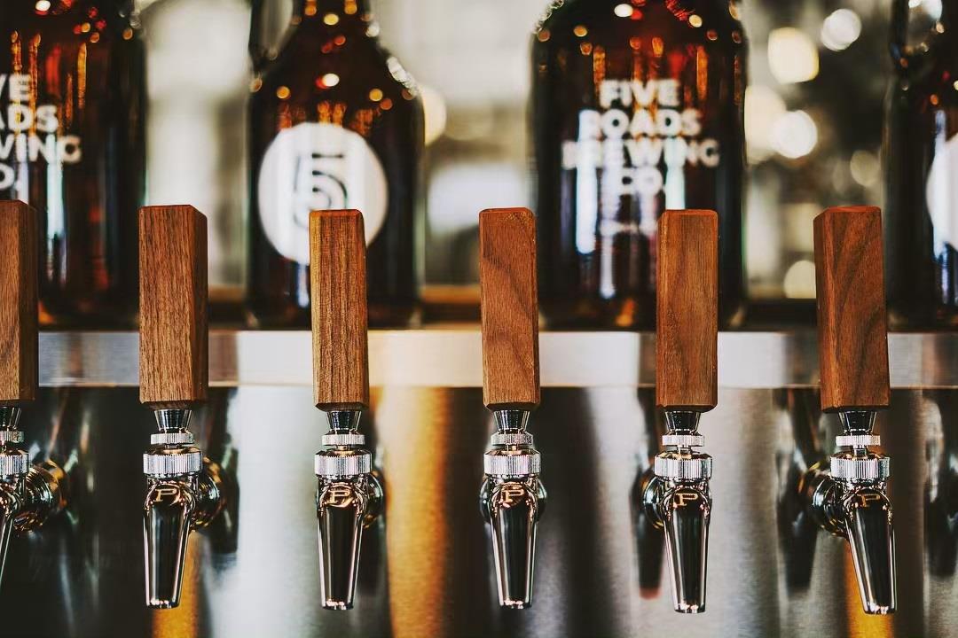 Row of beer taps with wooden handles in front of brown glass growlers labeled "Five Roads Brewing Co.