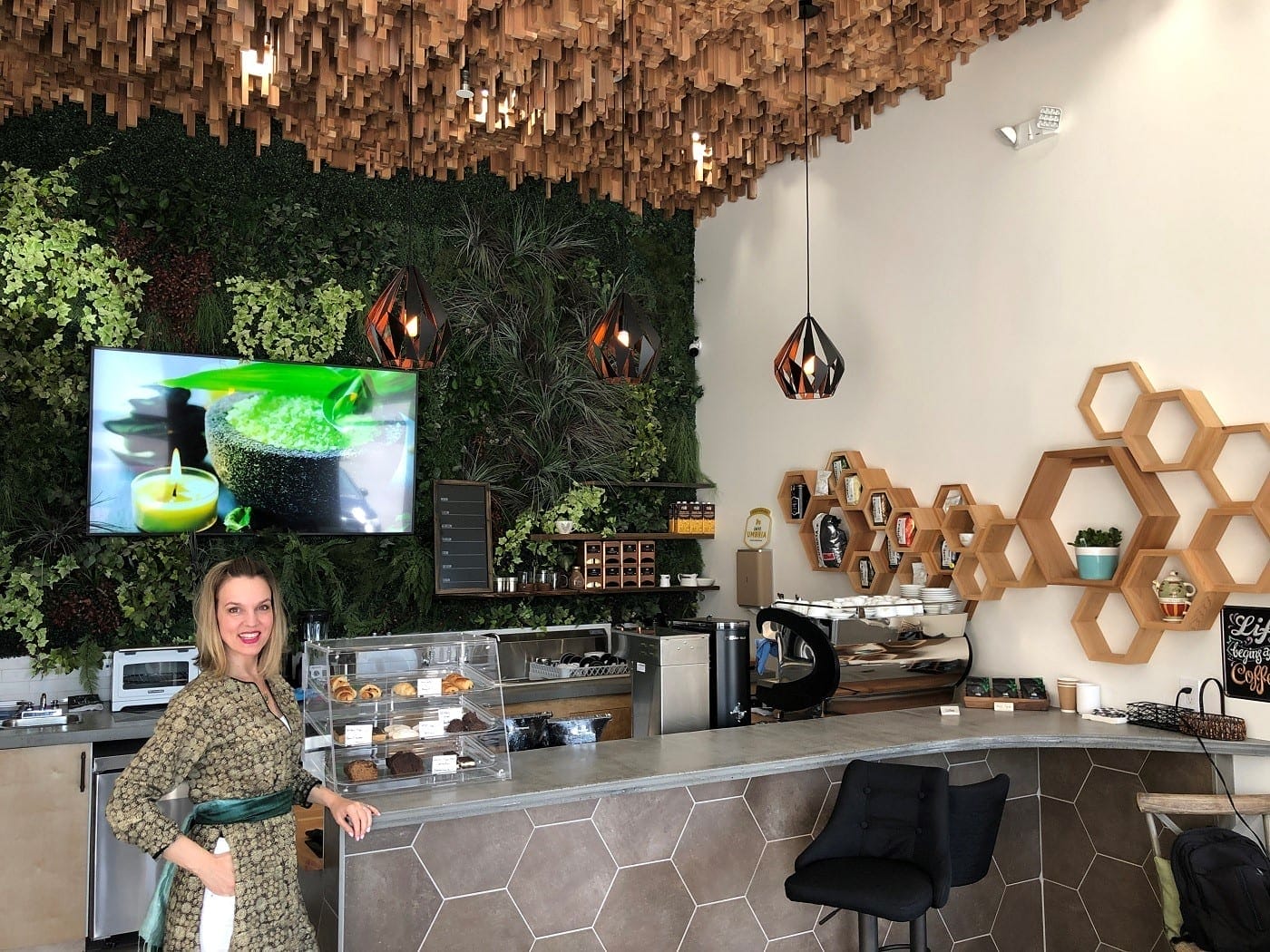 A woman stands at the counter of a modern café with geometric and plant-themed decor, a pastry display, and a digital screen on the wall behind her.