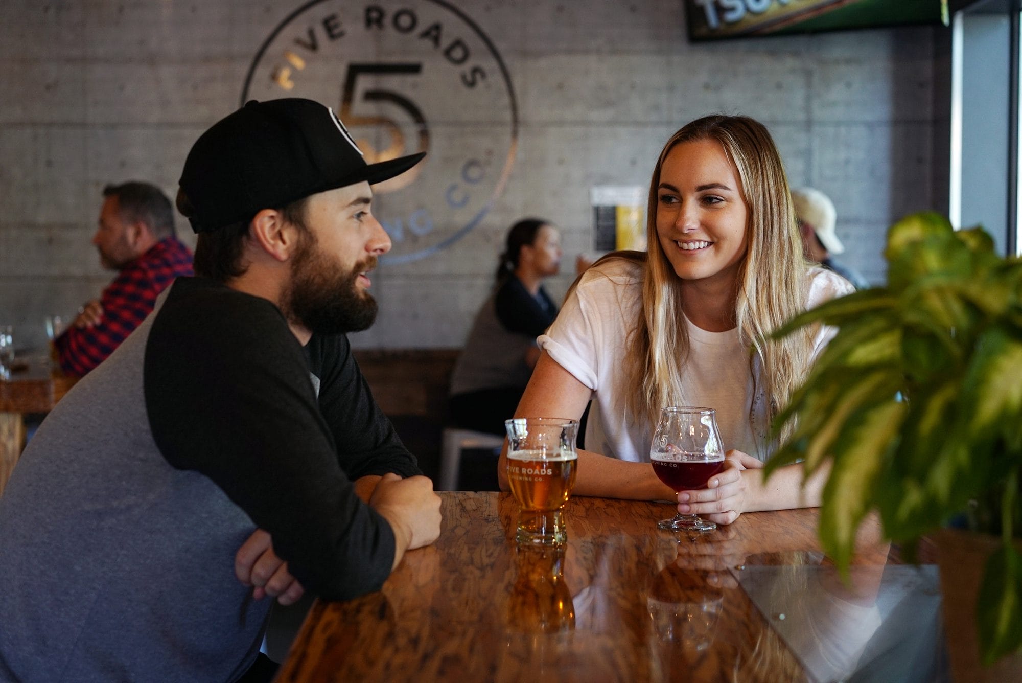 A man and woman sit at a wooden table in a bar, talking and smiling, with drinks in front of them. Other patrons are visible in the background.