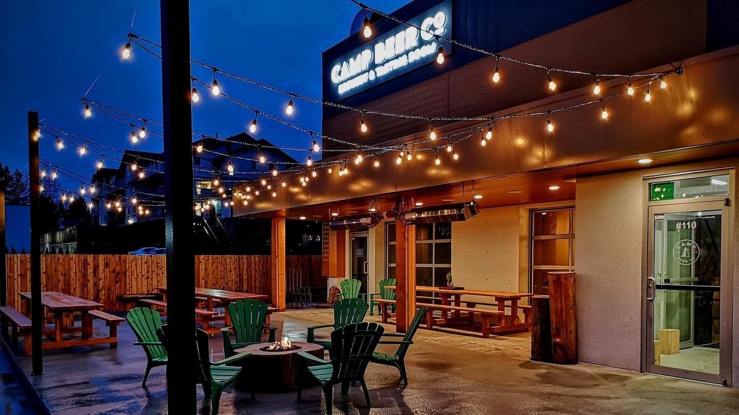 Outdoor patio at Camp Beer Co., lit with string lights, features green Adirondack chairs and wooden picnic tables on a wet concrete floor during dusk.