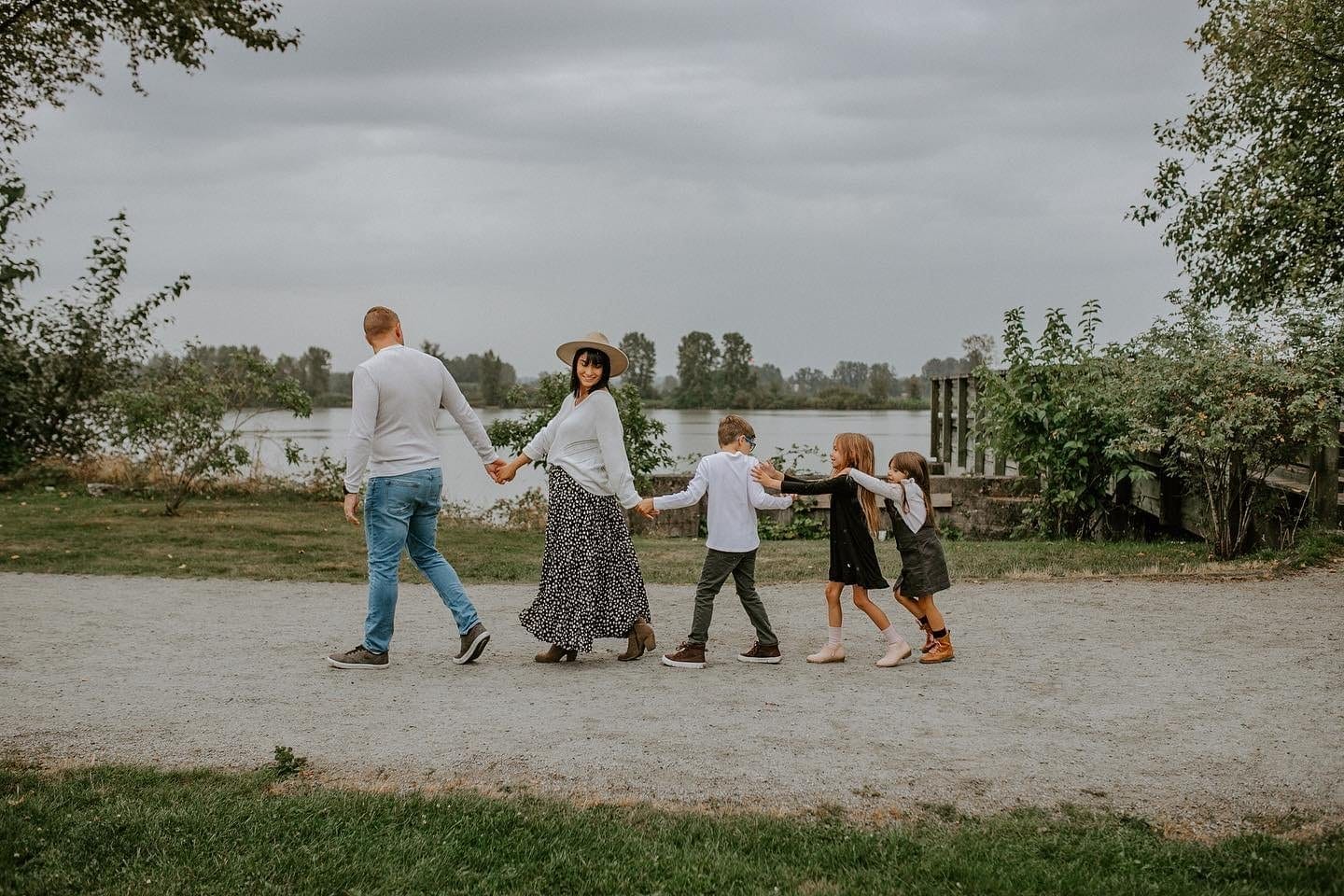 A family of five walks in a line holding hands on a gravel path near a river, with trees and cloudy sky in the background.