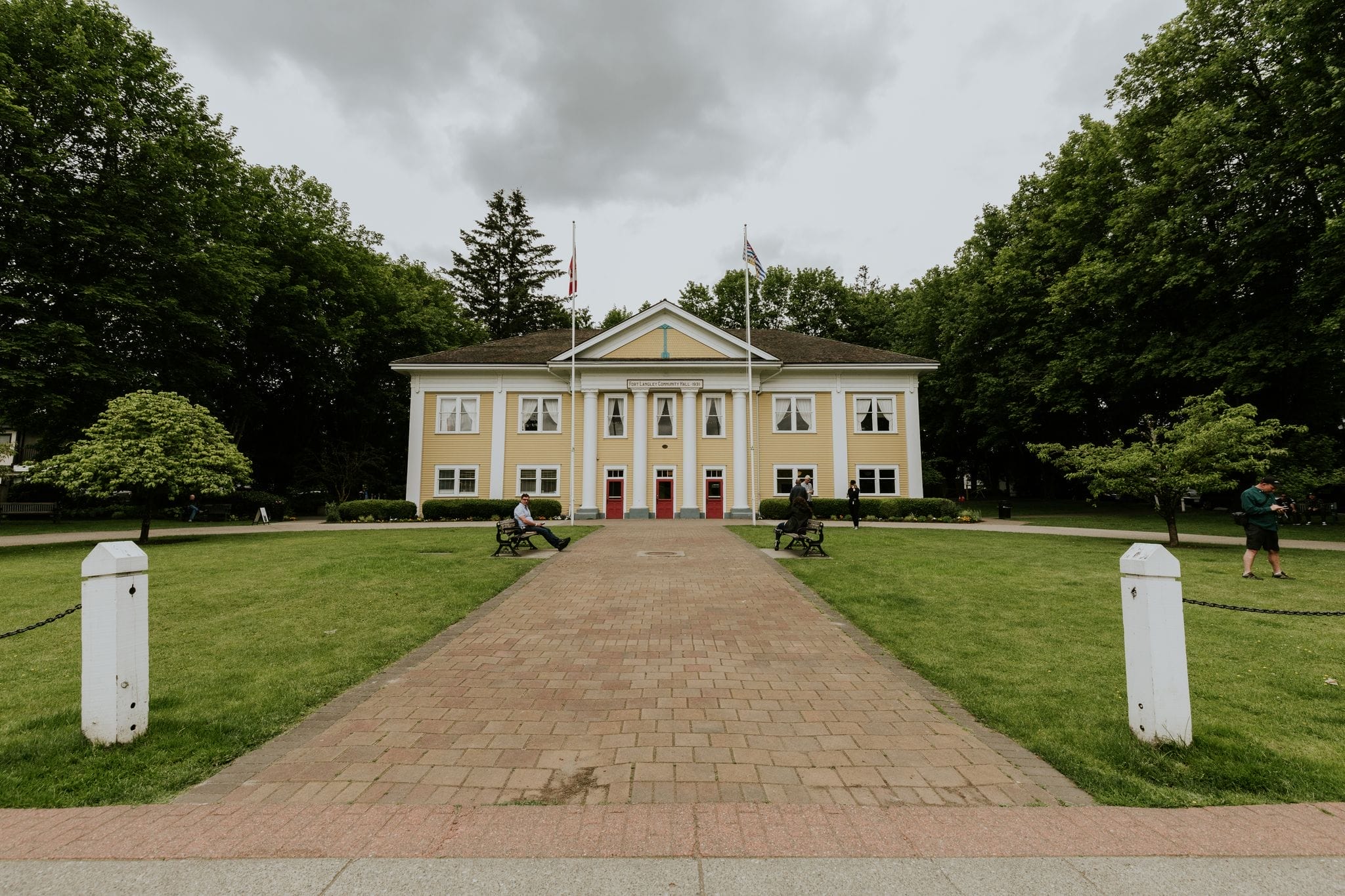 A yellow two-story building with three red doors stands at the end of a brick pathway, surrounded by green trees and lawn under a cloudy sky.