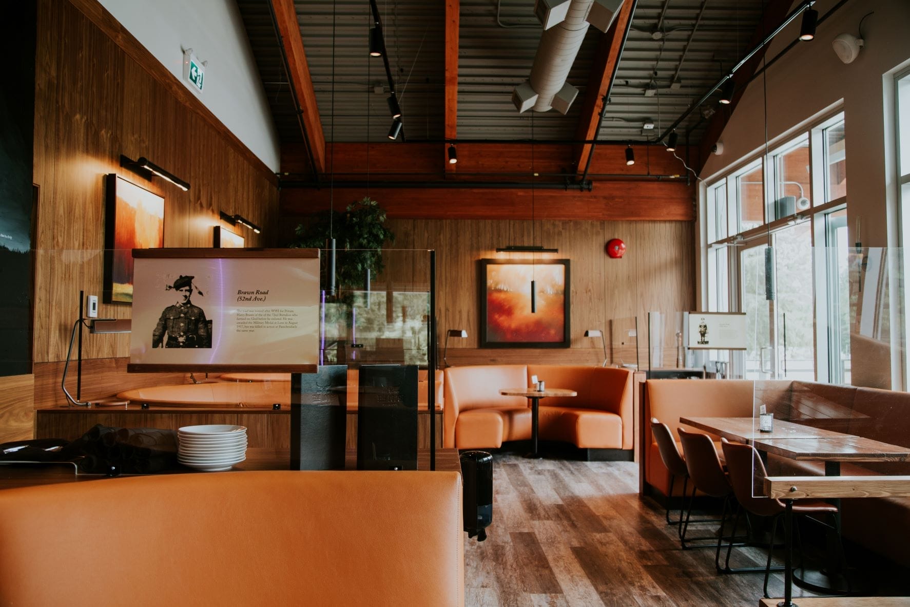 Modern restaurant interior with orange booths, wooden floor, large windows, and a TV screen displaying text and an image on the wall. Plexiglass dividers separate the seating areas.