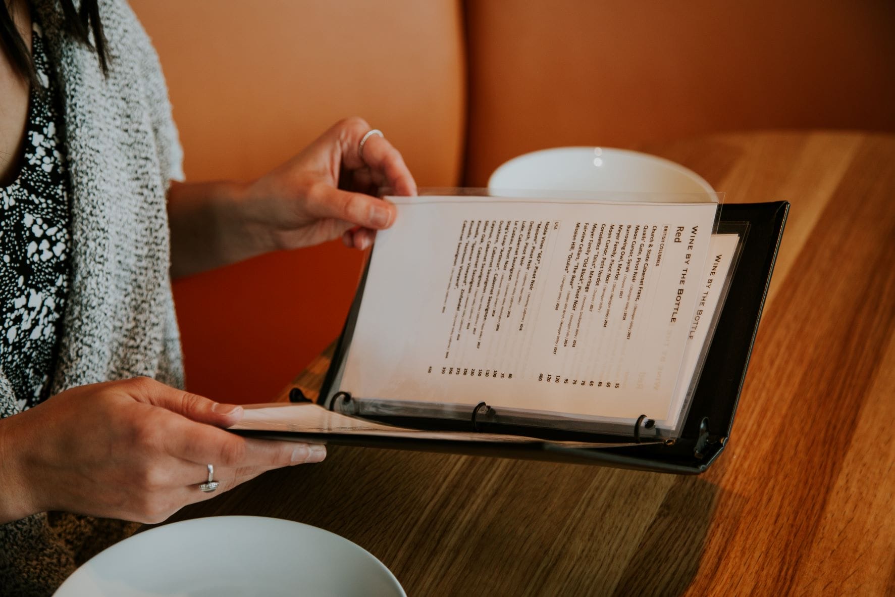 A person sits at a table holding and reading a restaurant menu in a plastic binder, with a white plate in front of them.