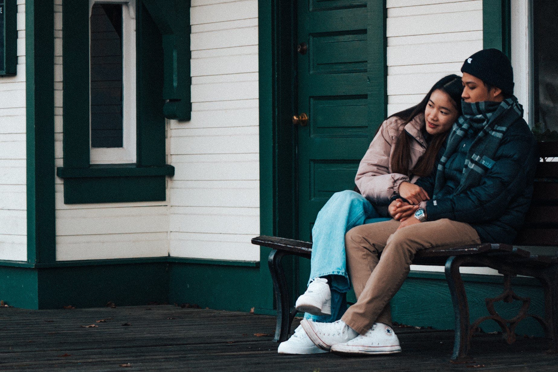 Two people sit closely together on a bench outside a green and white building, with one resting their head on the other's shoulder.