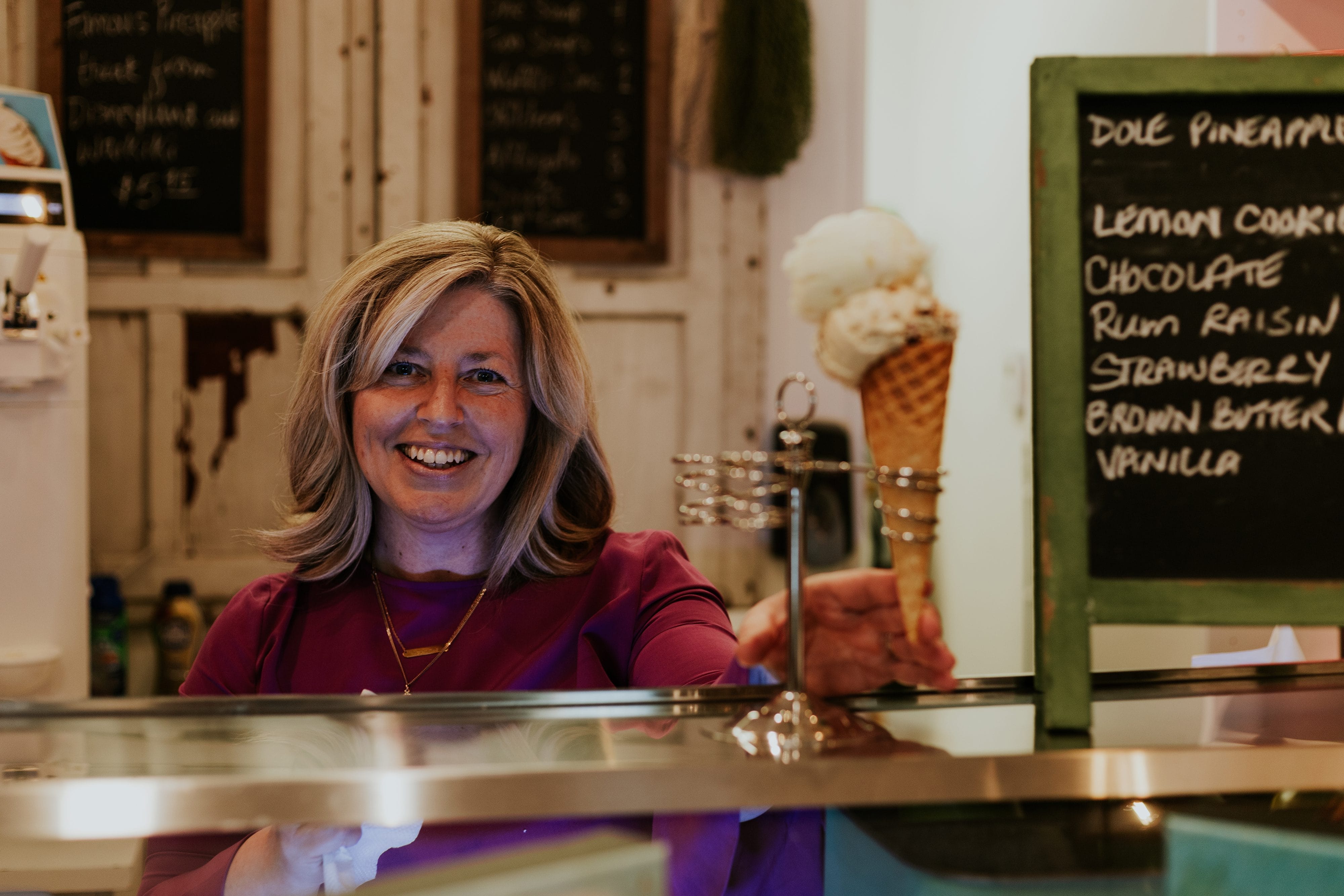 A woman in a purple top smiles behind an ice cream counter, with a waffle cone and a chalkboard menu listing ice cream flavors visible beside her.