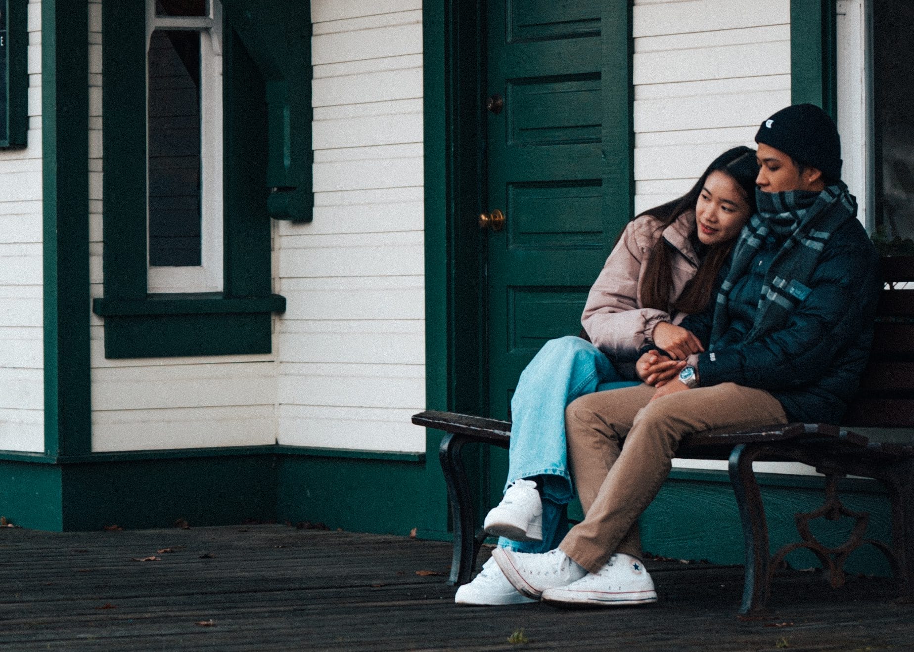 Two people sit closely together on a bench outside a building with green trim; one person rests their head on the other's shoulder, and they hold hands.