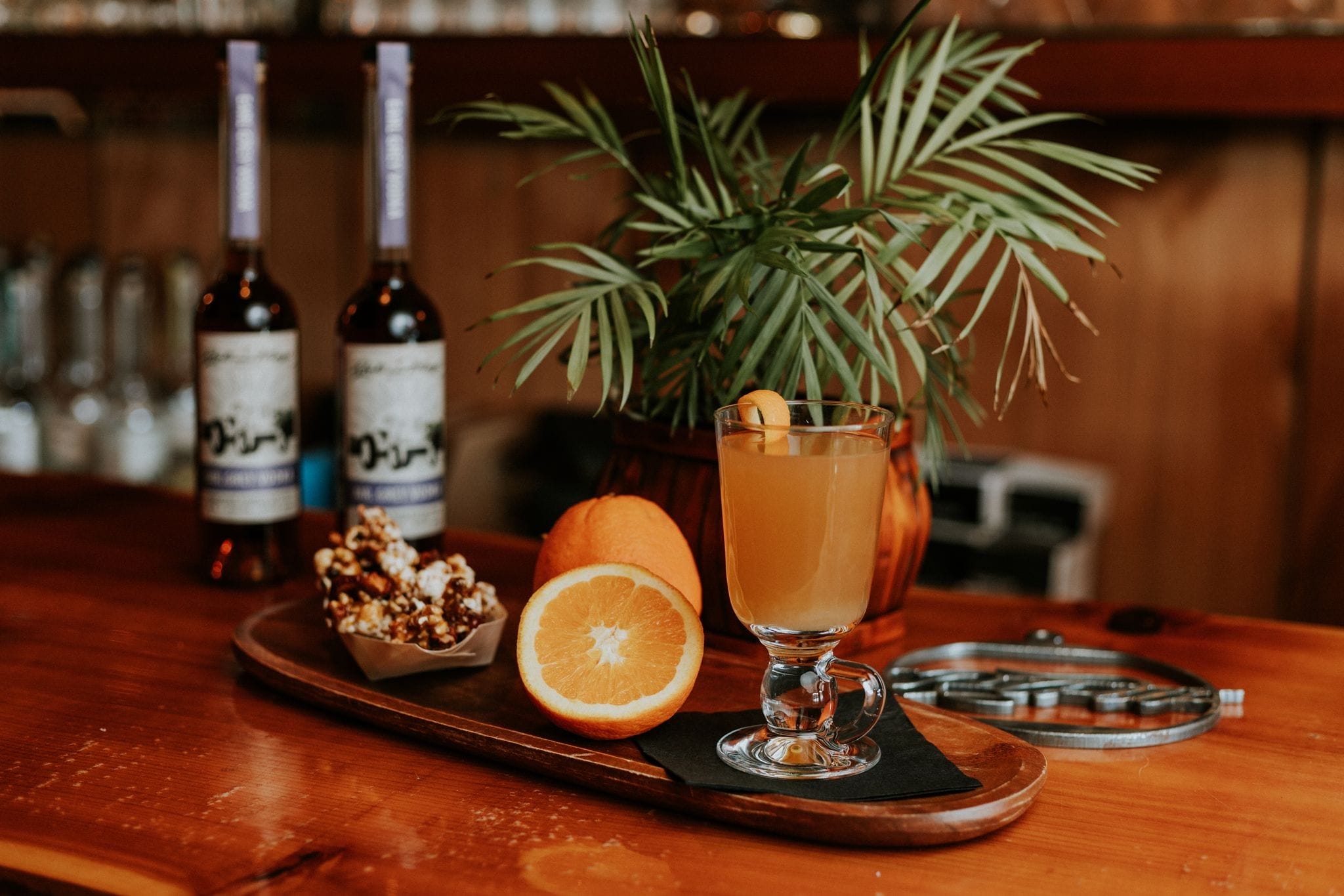 A glass of orange cocktail garnished with an orange peel sits on a wooden tray with popcorn, orange slices, and two bottles in the background, next to a potted plant.