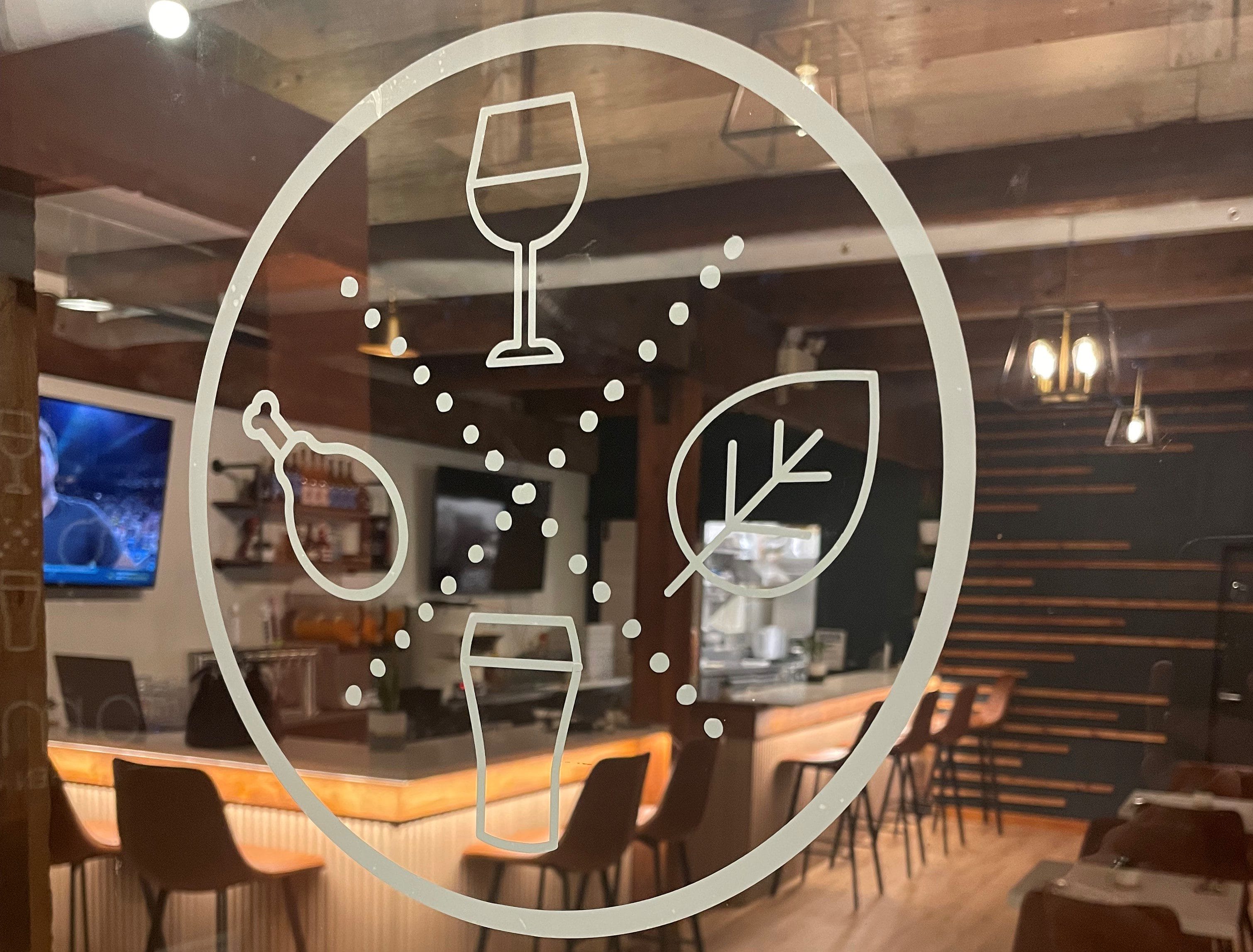 A transparent glass door with white line drawings of a wine glass, beer glass, meat leg, and leaf, with a bar and seating area visible inside.