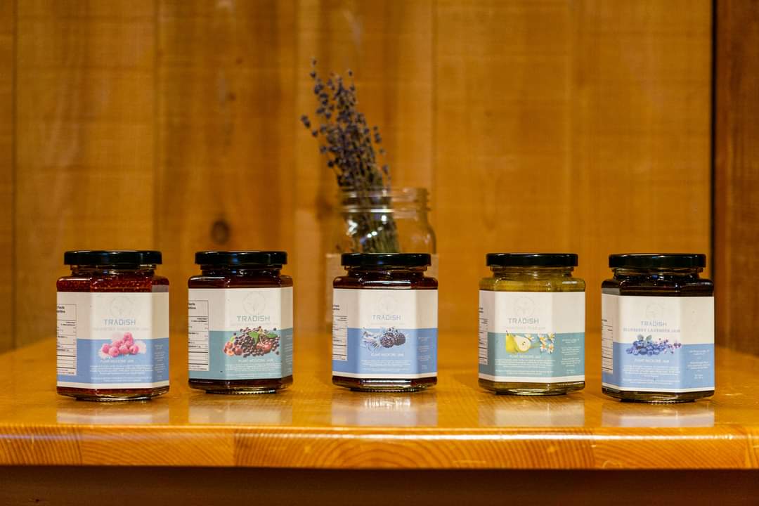Five jars of preserves with different fruit labels are arranged in a row on a wooden surface, with a glass jar of dried lavender in the background.
