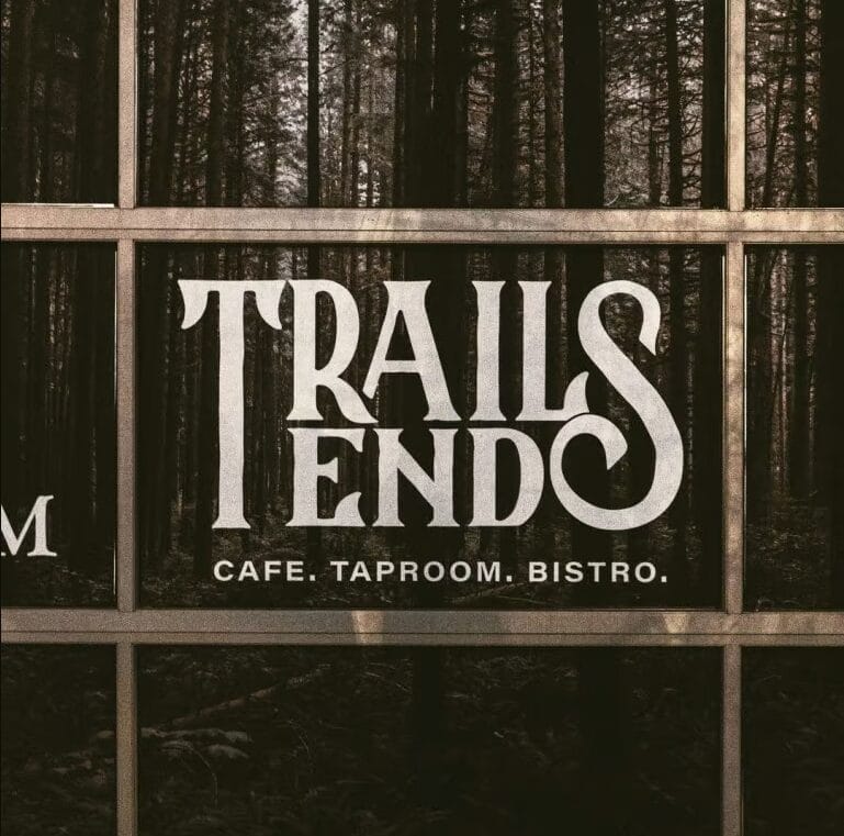 A window sign reads "TRAILS END CAFE. TAPROOM. BISTRO." with a forest reflected in the glass behind the text.