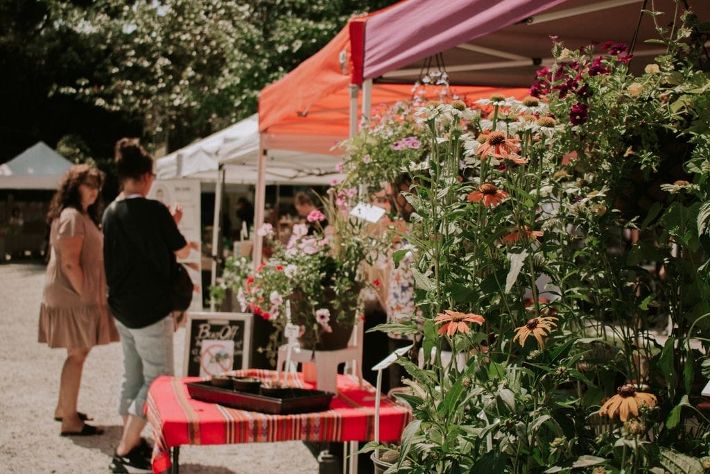 People at an outdoor market browse stalls under colorful tents, with potted flowers and plants displayed in the foreground on a sunny day.