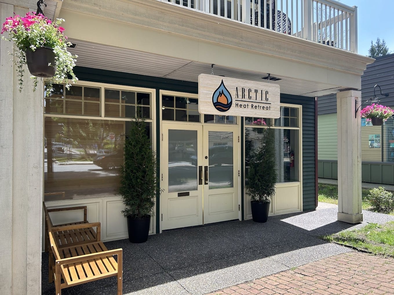 Street view of the entrance to Arctic Heat Retreat, featuring glass doors, a wooden bench, hanging flower baskets, and potted plants on either side of the doorway.