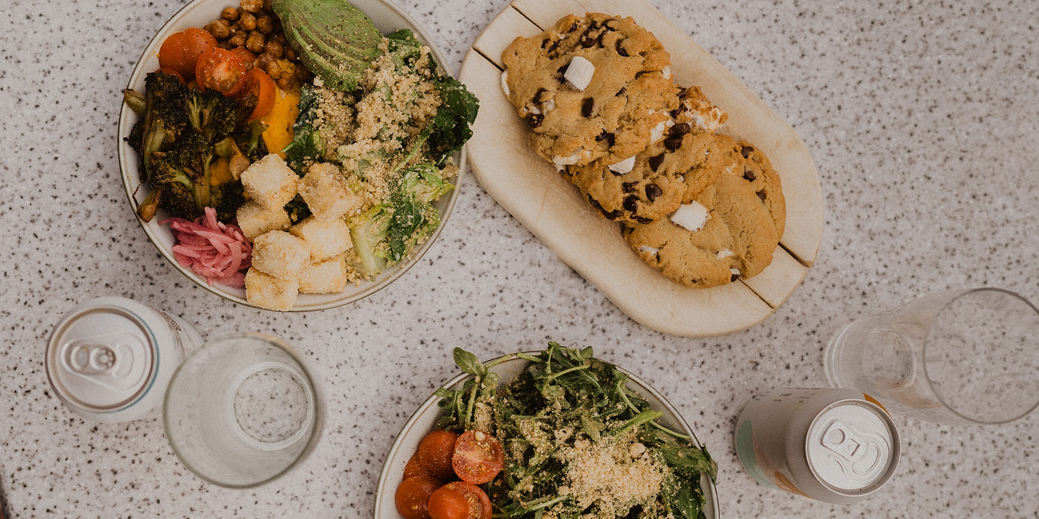 Two bowls of salad with vegetables and sprinkled toppings, a plate of chocolate chip cookies, two cans of sparkling water, and two glasses on a speckled tabletop.