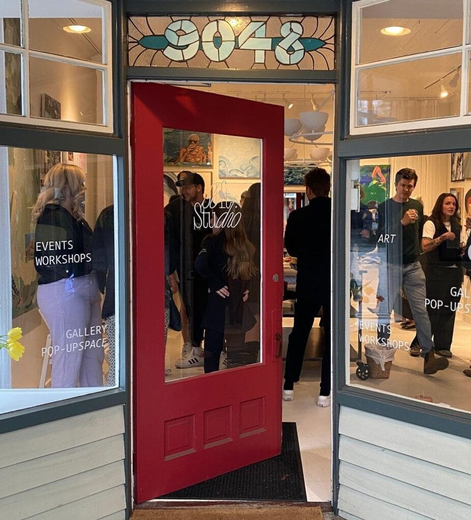 People gather inside an art gallery with a red door, stained glass window marked "9048," and signage for events, workshops, and gallery pop-ups visible on the glass.
