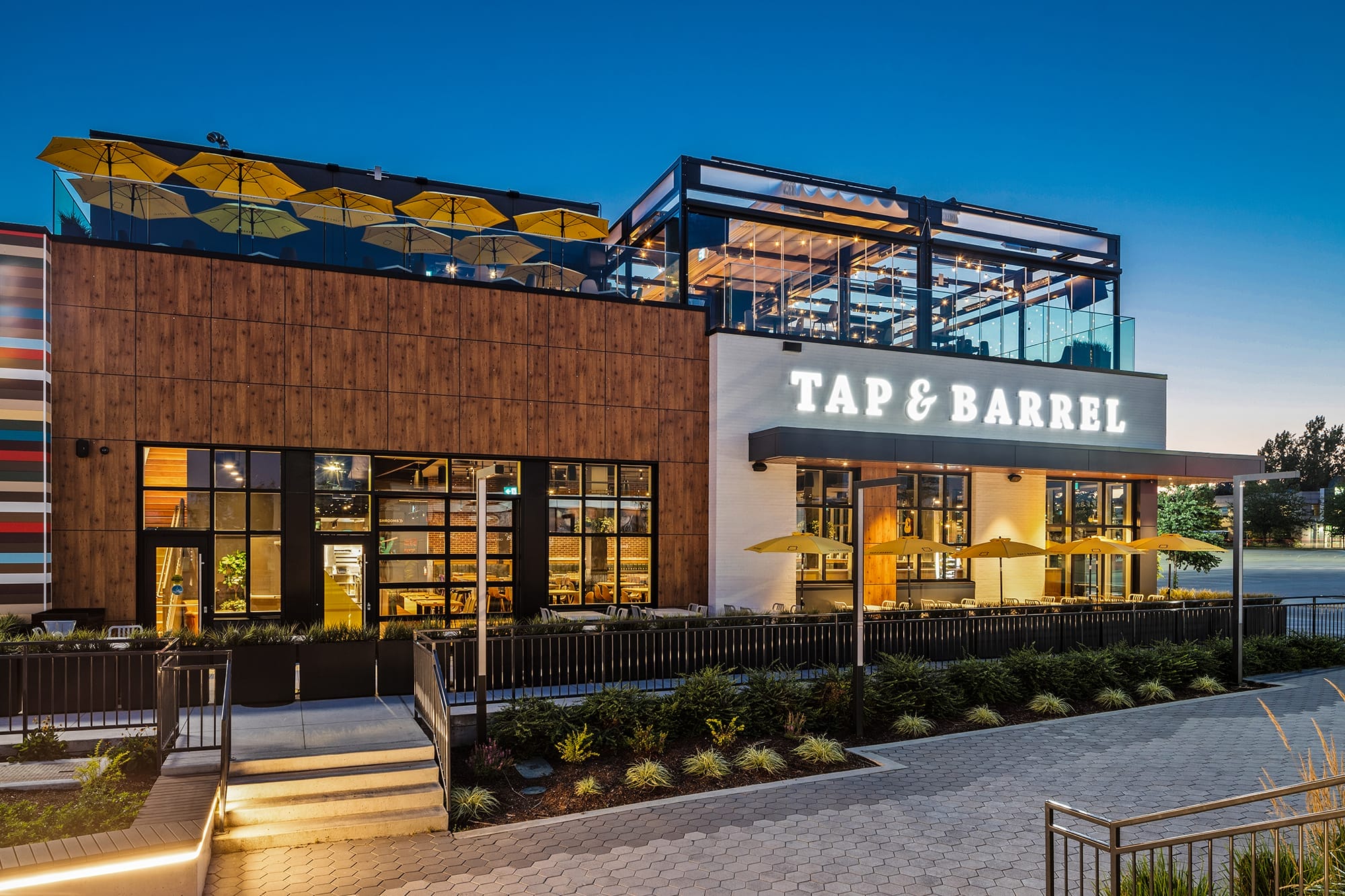 Modern two-story restaurant named Tap & Barrel with outdoor patio seating, large windows, and yellow umbrellas, photographed at dusk.