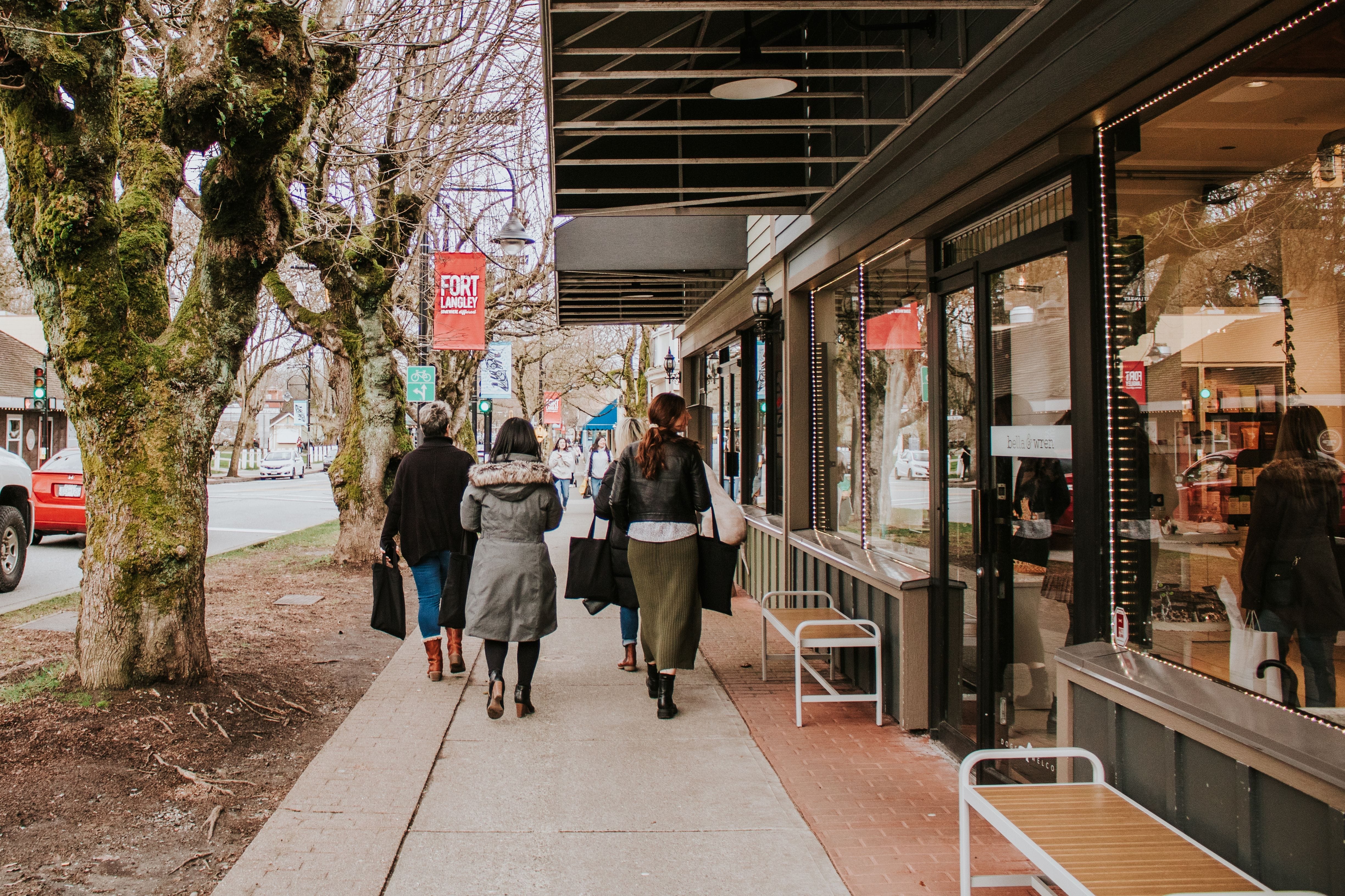 Five people walk along a sidewalk next to storefronts and trees on a cloudy day.