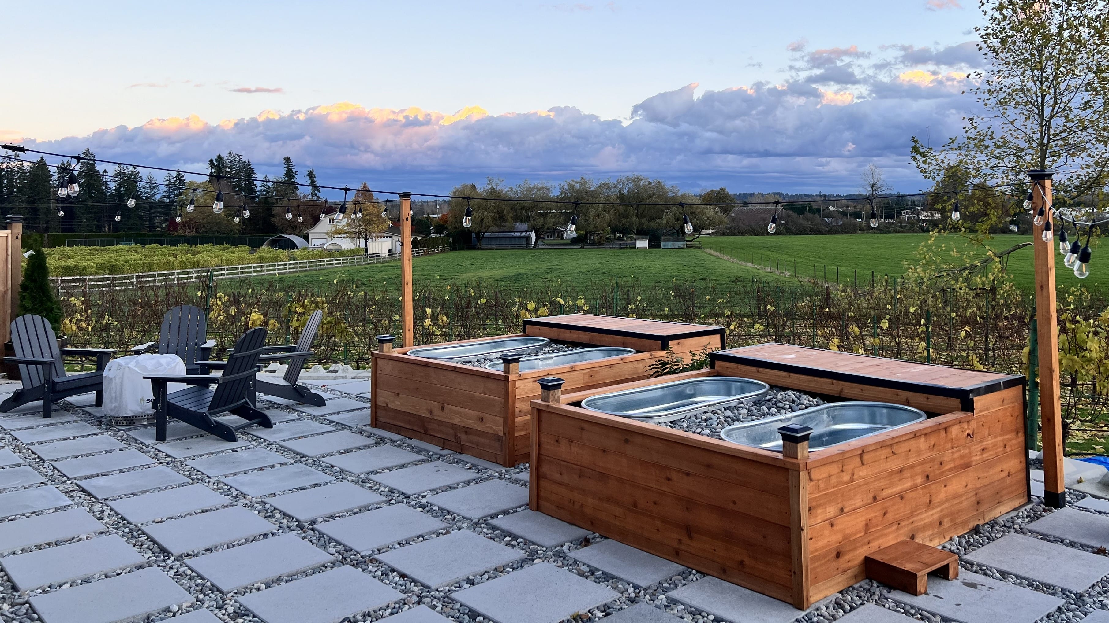 Two wooden hot tubs with metal interiors are on a stone patio, surrounded by Adirondack chairs, overlooking a green field and cloudy sky at sunset.