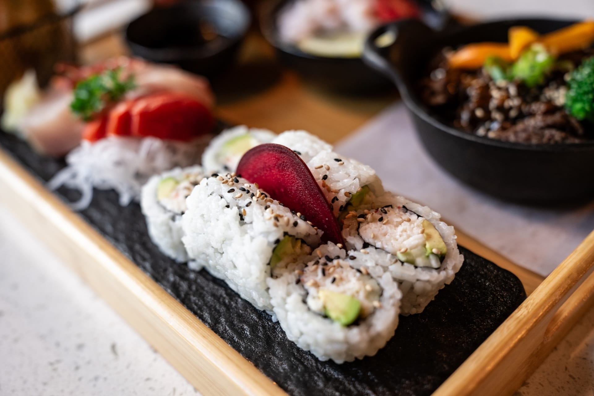 A close-up of sushi rolls with sesame seeds and avocado, garnished with a slice of pickled vegetable, served on a black rectangular plate with sashimi and other dishes in the background.