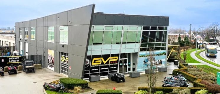 Modern two-story commercial building with large glass windows and “GVP” signage; several ATVs are parked outside near landscaped greenery and a curved road.