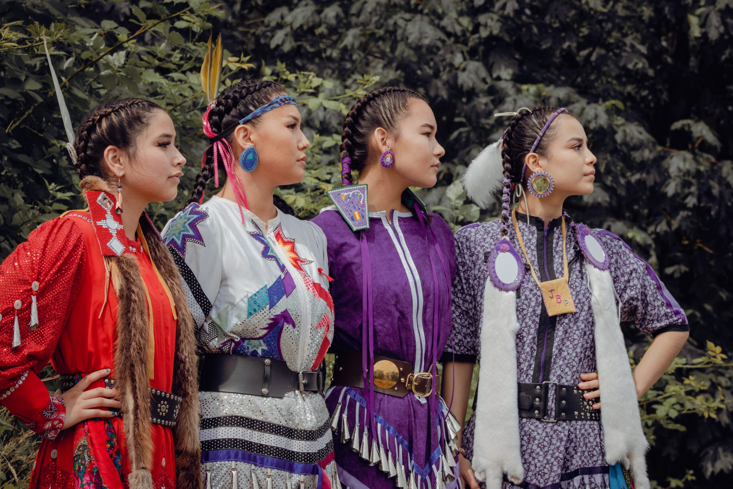 Four young women wearing colorful traditional regalia stand side by side outdoors, facing right, with trees in the background.