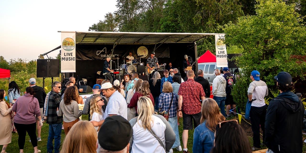 A crowd stands outdoors watching a live band perform on a stage surrounded by trees and tents during the day.