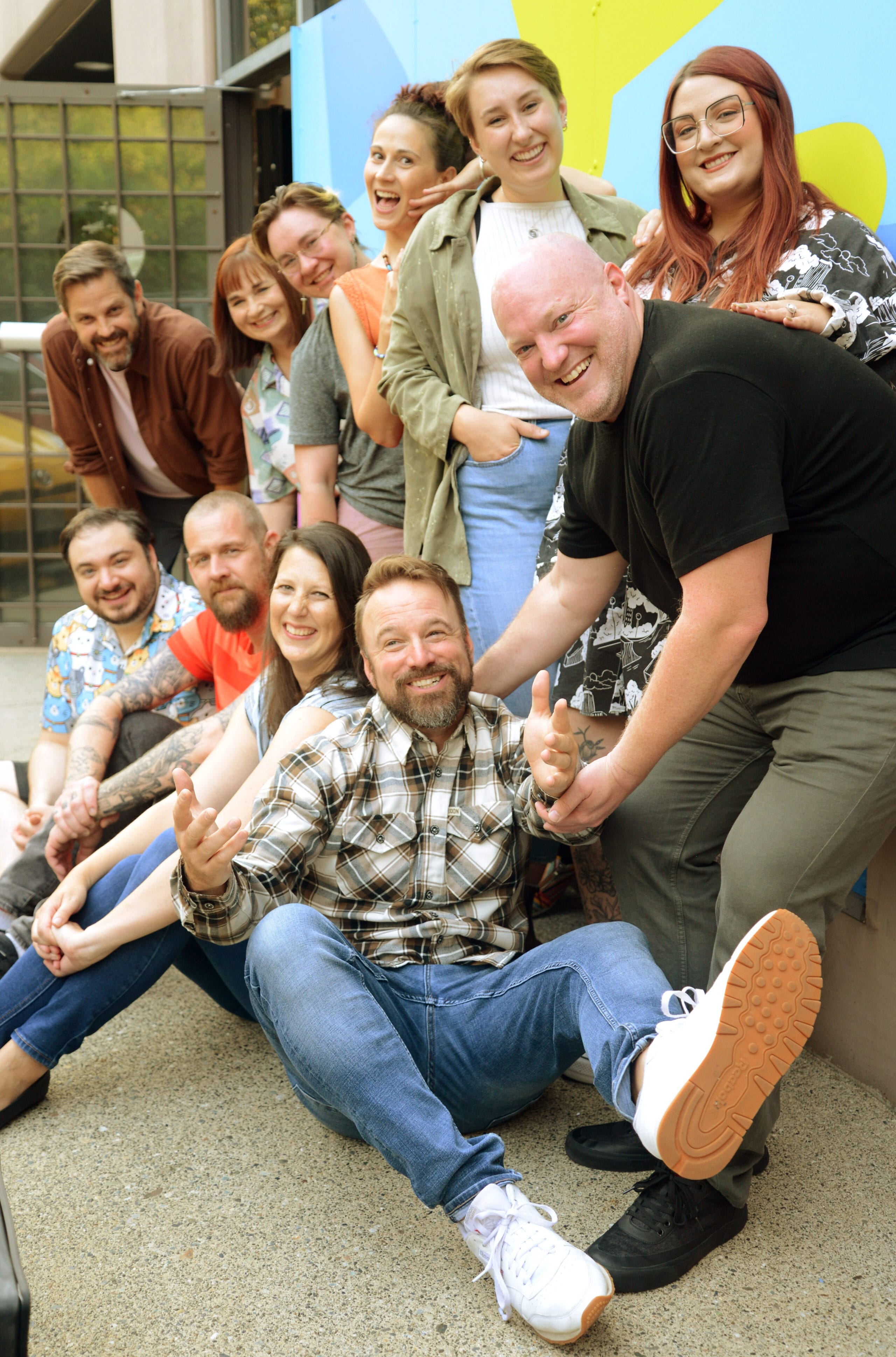 A group of twelve adults pose closely together outdoors, smiling and laughing for the camera.
